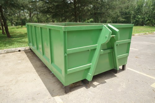 A large, rectangular green skip bin with reinforced metal sides and a smooth finish, positioned on a paved surface in an outdoor area with trees and grass visible in the background. The bin is elevated slightly on wooden sleepers, allowing clearance underneath. Its front side features a sturdy, hinged metal latch and a wide, open top for easy loading of bulky rubbish or waste materials. The surrounding environment suggests an open space typical of commercial or waste management sites, relevant to rubbish removal services offered by Commercial Waste Chigwell. The sunlight highlights the fresh, vibrant green colour of the skip, contrasting with the natural greenery behind it. The image captures the clean, well-maintained appearance of the bin, emphasizing its suitability for collecting general waste or construction debris in a suburban or semi-rural setting consistent with UK waste disposal practices in the Chigwell area.