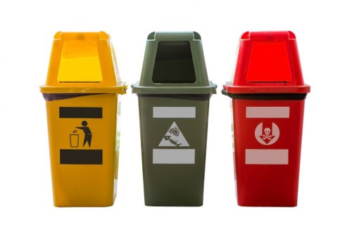 A digital illustration of three colour-coded waste bins typically used for rubbish disposal. The first bin on the left is yellow with a black graphic depicting a person disposing of waste into a container, suggesting general waste or packaging. The middle bin is green with a white symbol illustrating food or organic waste, indicating compost or biodegradable rubbish. The third bin on the right is red with a white hazard symbol symbolising hazardous or clinical waste. The bins are positioned side by side in a well-lit setting, with their open lids revealing empty interiors, each made of durable plastic with smooth, glossy surfaces. The background is plain and neutral, focusing attention on the bins' features, which are common for waste management services in urban or suburban environments, including towns like Chigwell. These bins are associated with commercial waste services such as those provided by Commercial Waste Chigwell, highlighting their role in segregating different types of rubbish for proper disposal. The image emphasizes clear colour coding and graphic symbols essential for effective rubbish sorting and collection.