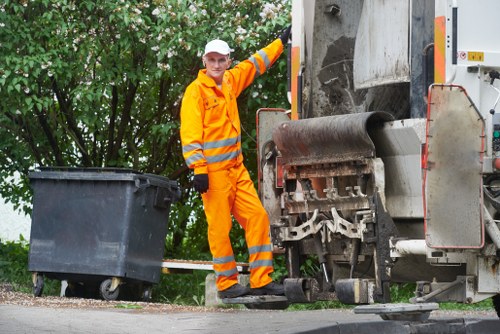 A waste management worker dressed in high-visibility orange overalls with reflective stripes, standing next to a commercial rubbish collection vehicle on a residential street. The worker is smiling and holding a large black wheeled rubbish bin, positioned on the pavement near the rear of the vehicle. The waste truck, which features a metallic side panel and a rotating compacting mechanism, occupies the right side of the image and appears to be in preparation for or engaged in rubbish collection. In the background, there are green leafy trees and bushes, indicating a suburban environment. The scene is well-lit, suggesting daytime, and the pavement shows typical roadway features. This setting demonstrates appropriate rubbish removal service procedures, relevant to the locality served by Commercial Waste Chigwell in Essex, with a focus on efficient waste collection on a quiet street aligned with local residential waste management practices.