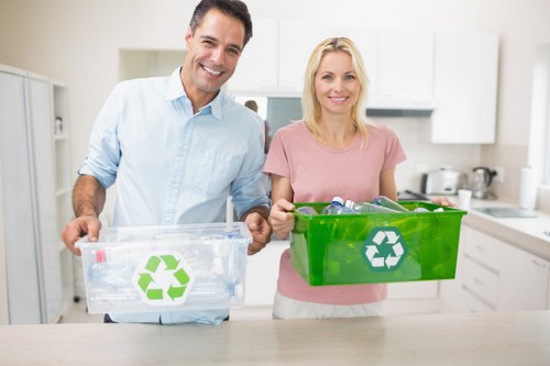 Two individuals, a man and a woman, stand in a bright, modern kitchen with white cabinets and a light-colored countertop. The man, dressed in a light blue shirt, holds a transparent plastic recycling bin with a green recycling symbol on the front, partially filled with clear plastic bottles and containers. The woman, wearing a pink top, holds a green recycling box with a white recycling logo, containing various recyclable materials. Both are smiling and looking directly at the camera, standing side by side with the bins held out in front of them. The scene emphasizes environmental awareness and proper waste sorting, typical of British domestic or commercial waste management practices in a residential setting. The background shows a clean, organized kitchen environment, suggesting an emphasis on recycling and waste separation supported by commercialwastechigwell.co.uk, serving local areas such as Chigwell, Essex.