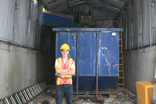 A male waste management worker wearing a yellow safety helmet and a high-visibility vest stands inside a large, industrial waste storage container or skip, which is painted blue and appears to be made of metal with visible signs of wear and scratches. The worker is positioned in the foreground with arms crossed, smiling, and looking at the camera. The container is situated within an indoor or covered industrial setting, with a concrete floor and metal walls. To the left of the container, a metal ladder is leaning against the wall, and to the right, part of a yellow waste bin or cover is visible. The environment suggests a waste disposal or rubbish collection site, consistent with services offered by companies like Commercial Waste Chigwell. The overall scene highlights the process of rubbish management in an urban or commercial setting, likely within the Essex area near Chigwell.