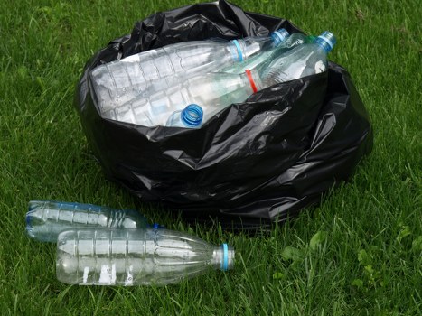 An open black rubbish bag filled with several transparent plastic bottles, some with blue caps and one with a red mark, is placed on a grassy outdoor area. The bag is partially tied and bulging with the bottles, which vary in position, with some protruding from the top. A few bottles are lying on the grass nearby, indicating they have recently been discarded or removed. The surrounding environment features lush green grass, suggesting a garden or park space, and the lighting appears natural, with soft shadows. This scene depicts typical waste disposal of plastic bottles and aligns with rubbish removal services that handle household or commercial waste in areas around Chigwell, Essex. Commercial Waste Chigwell offers environmentally responsible waste collection options, as demonstrated by the tossed plastic bottles in this outdoor setting, reflecting the importance of proper disposal and recycling practices within the local community.